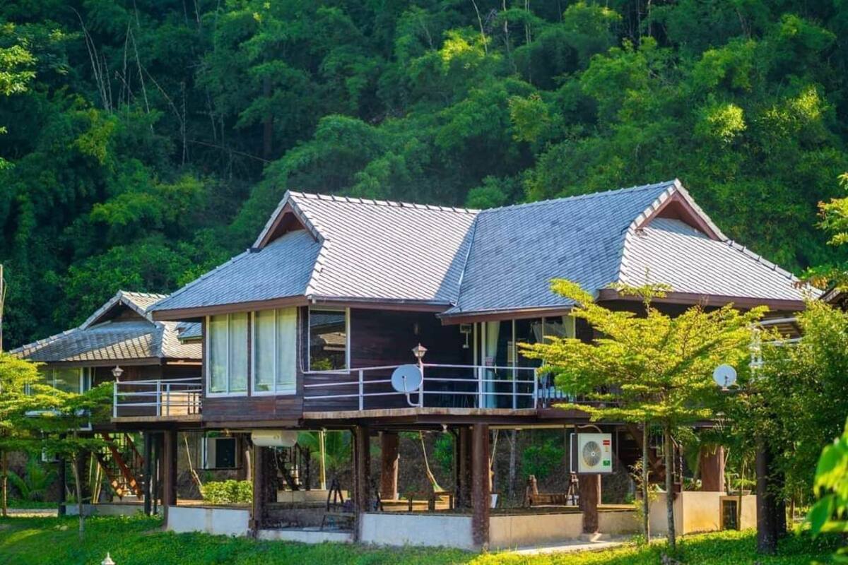 A traditional wooden house with a balcony, surrounded by lush greenery.