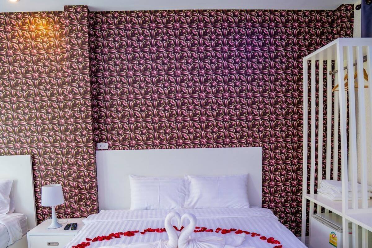 A hotel room with a bed, white bedding, a headboard, a nightstand, a lamp, a crib, and a wall with a floral pattern.