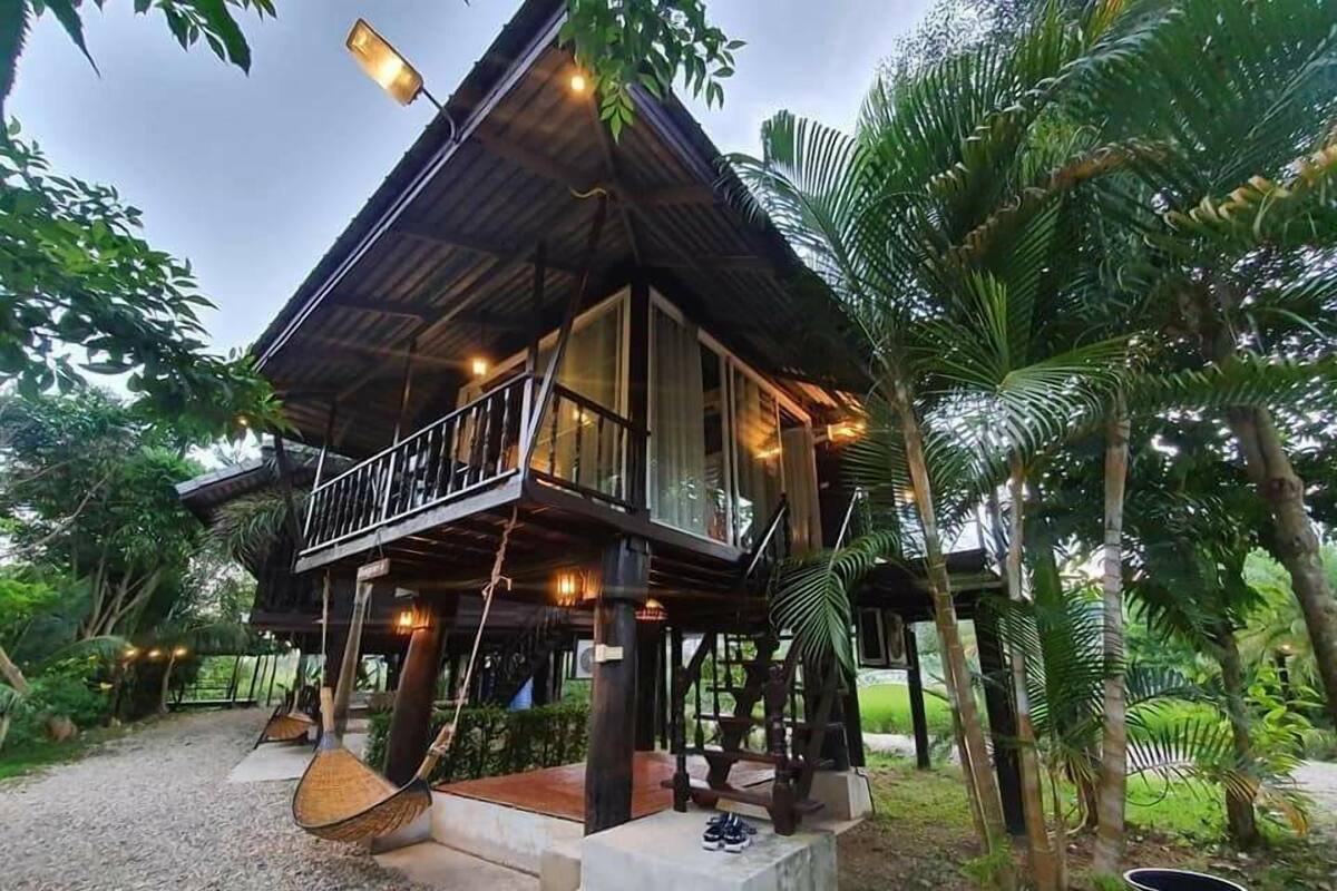 A two-story wooden house with a balcony, surrounded by tropical vegetation.