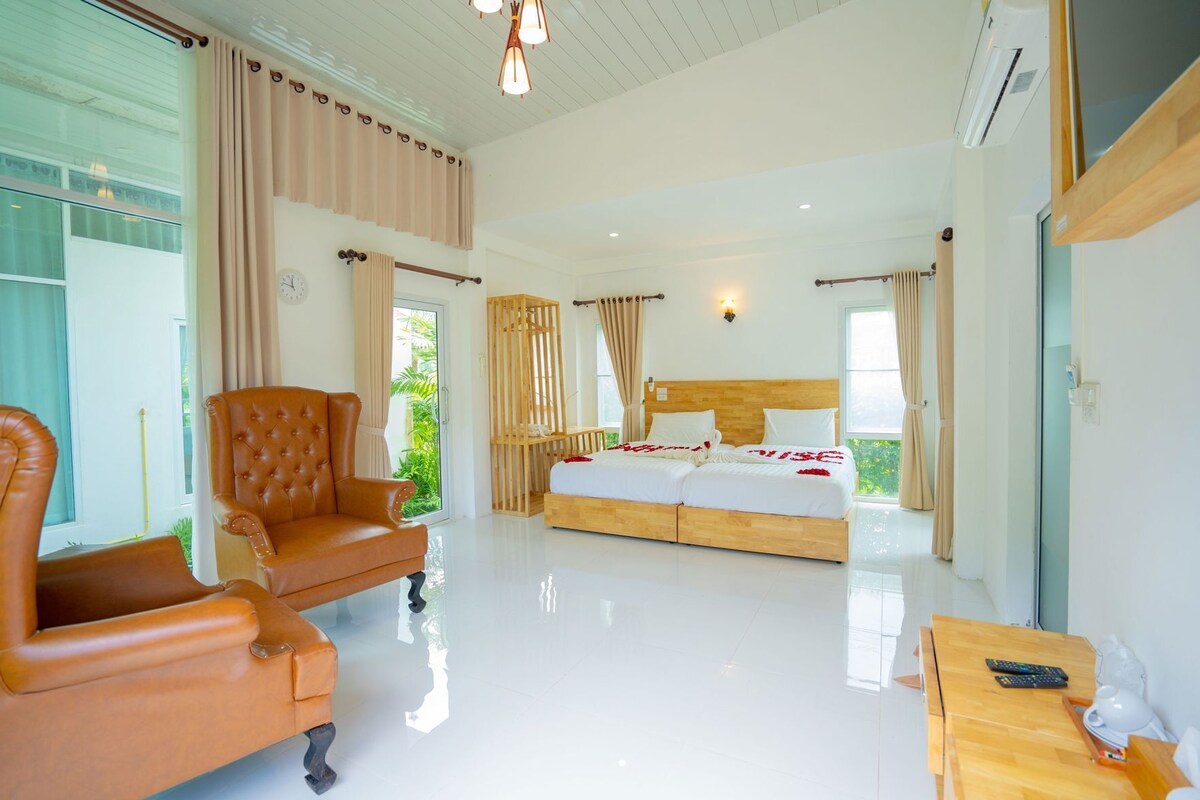 A spacious bedroom with a large bed, a leather armchair, a wooden bedside table, and a view of greenery outside.