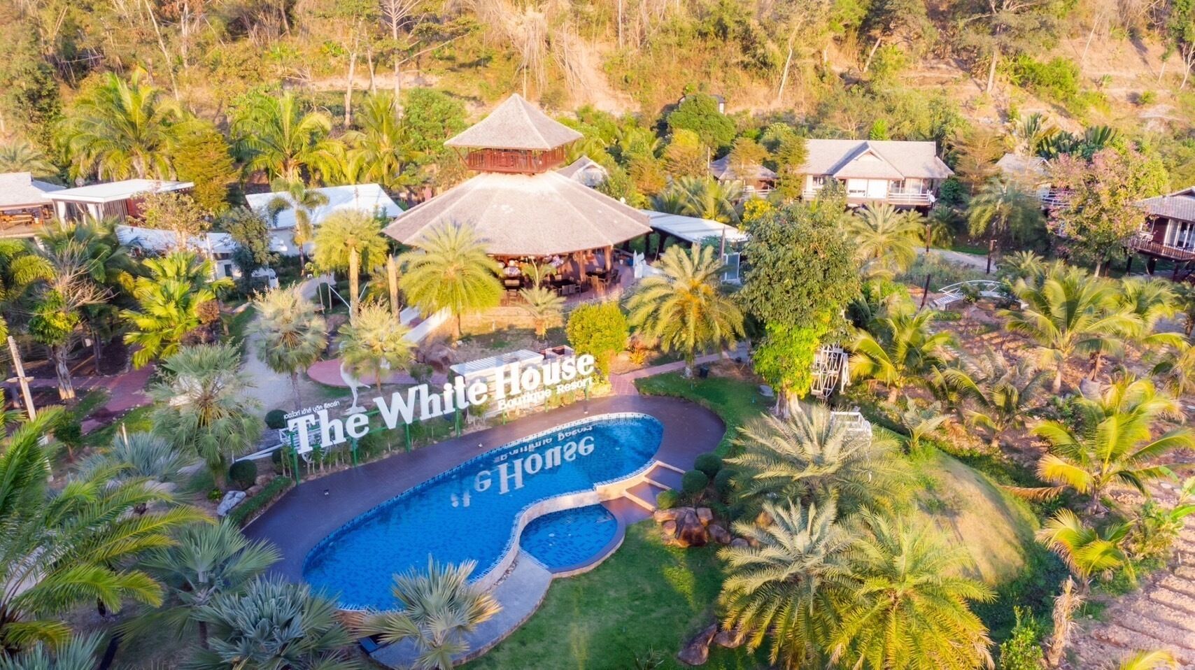 An aerial view of The White House Boutique Resort with a swimming pool, palm trees, and a thatched-roof building.