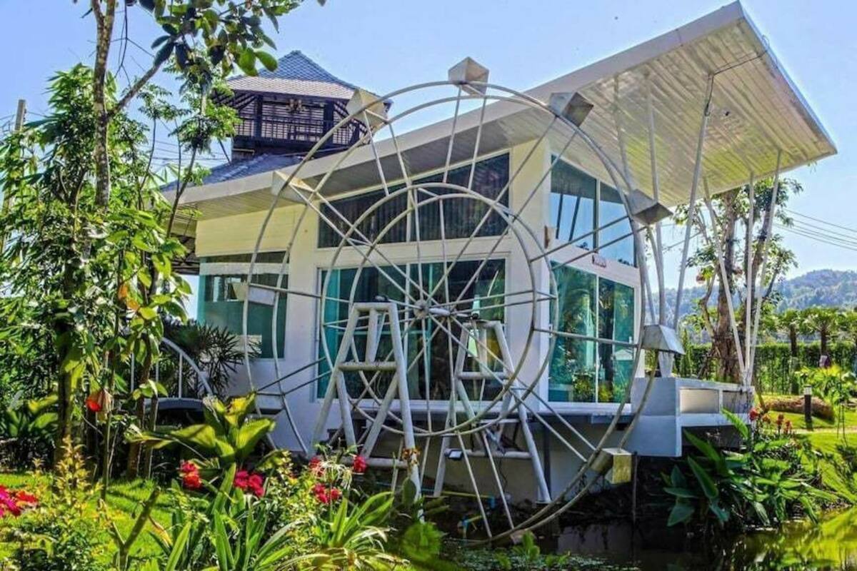 A modern house with a large circular window structure, surrounded by lush greenery and a pond.