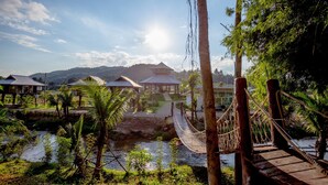 A riverside view with a wooden bridge, palm trees, and a building with a thatched roof. - The White House Boutique Chiangrai (Chiang Rai)
