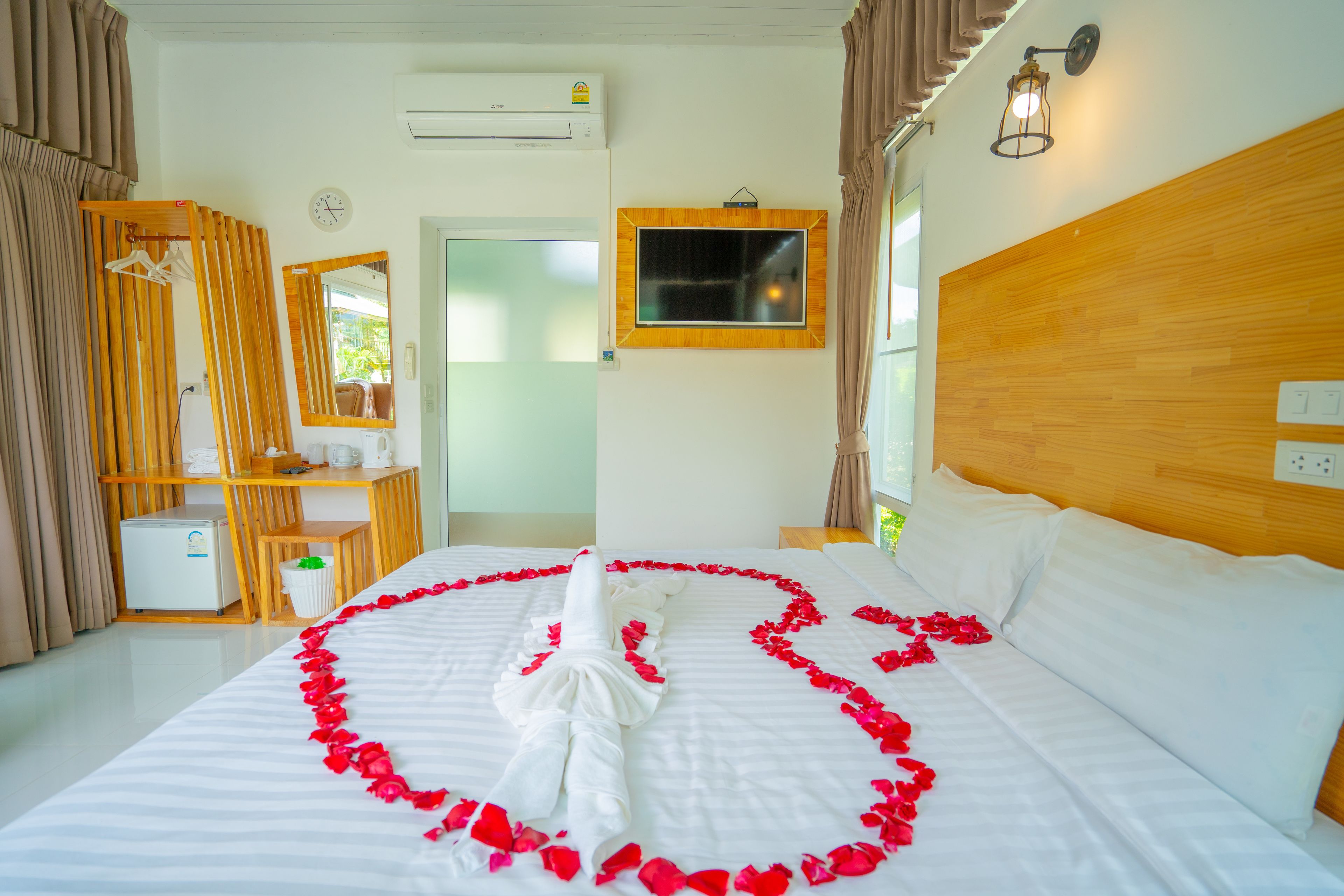A hotel room with a neatly made bed, a wooden headboard, a small desk, a mirror, and an air conditioning unit.