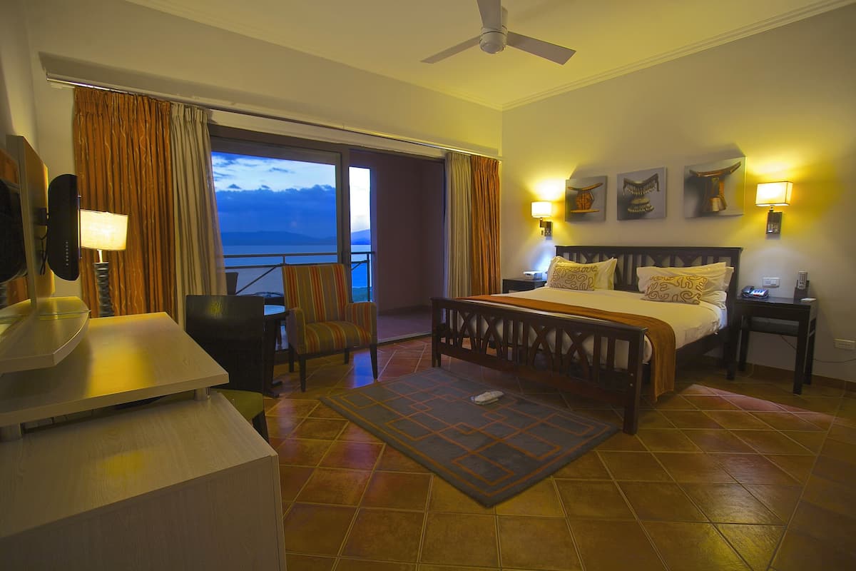 A hotel room with a bed, a desk, a chair, a balcony with curtains, and a view of the sky.