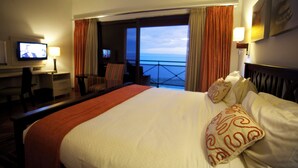 A hotel room with a bed, a desk, a television, and a view of the ocean.