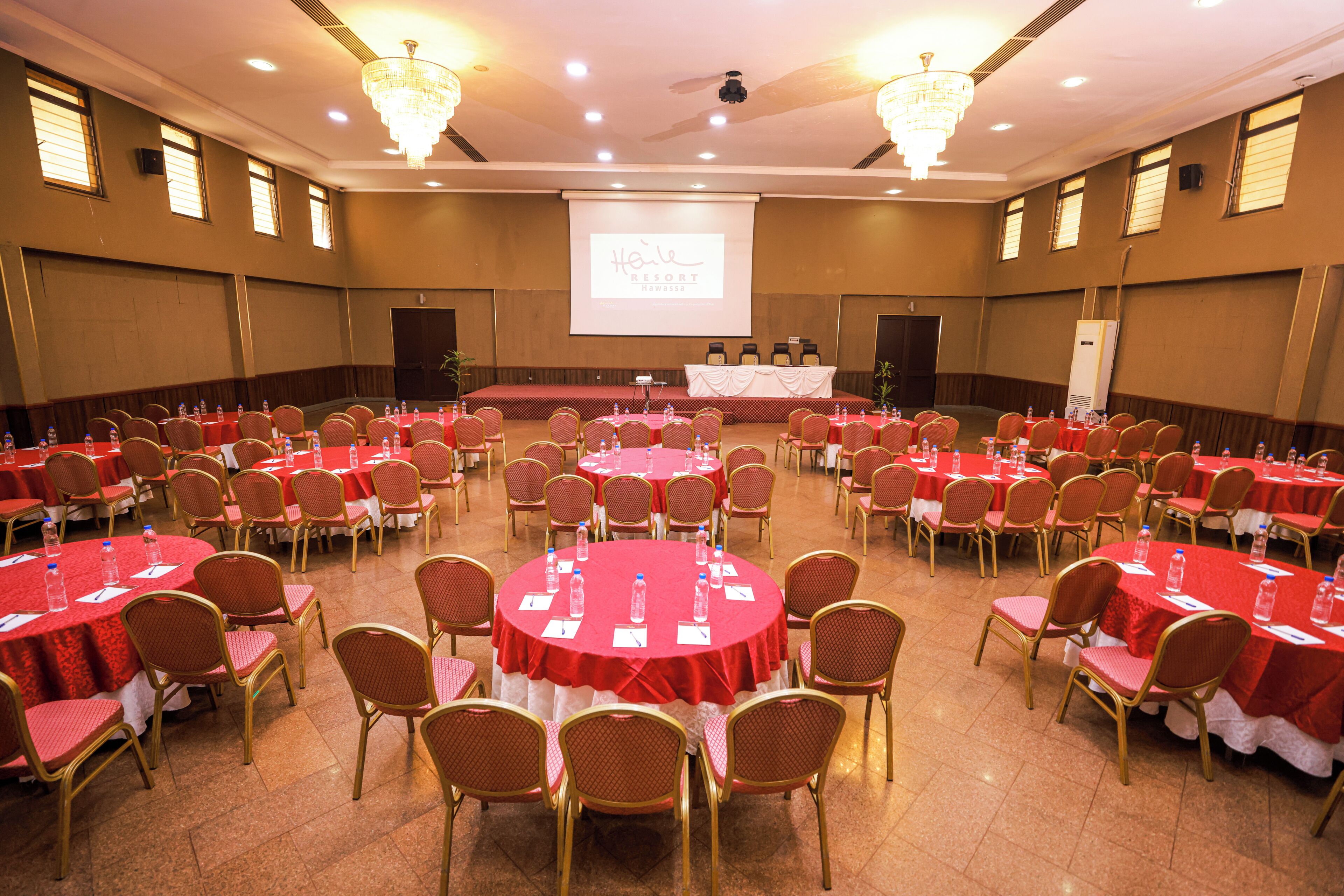A conference room with round tables, chairs, and a stage with a presentation screen.