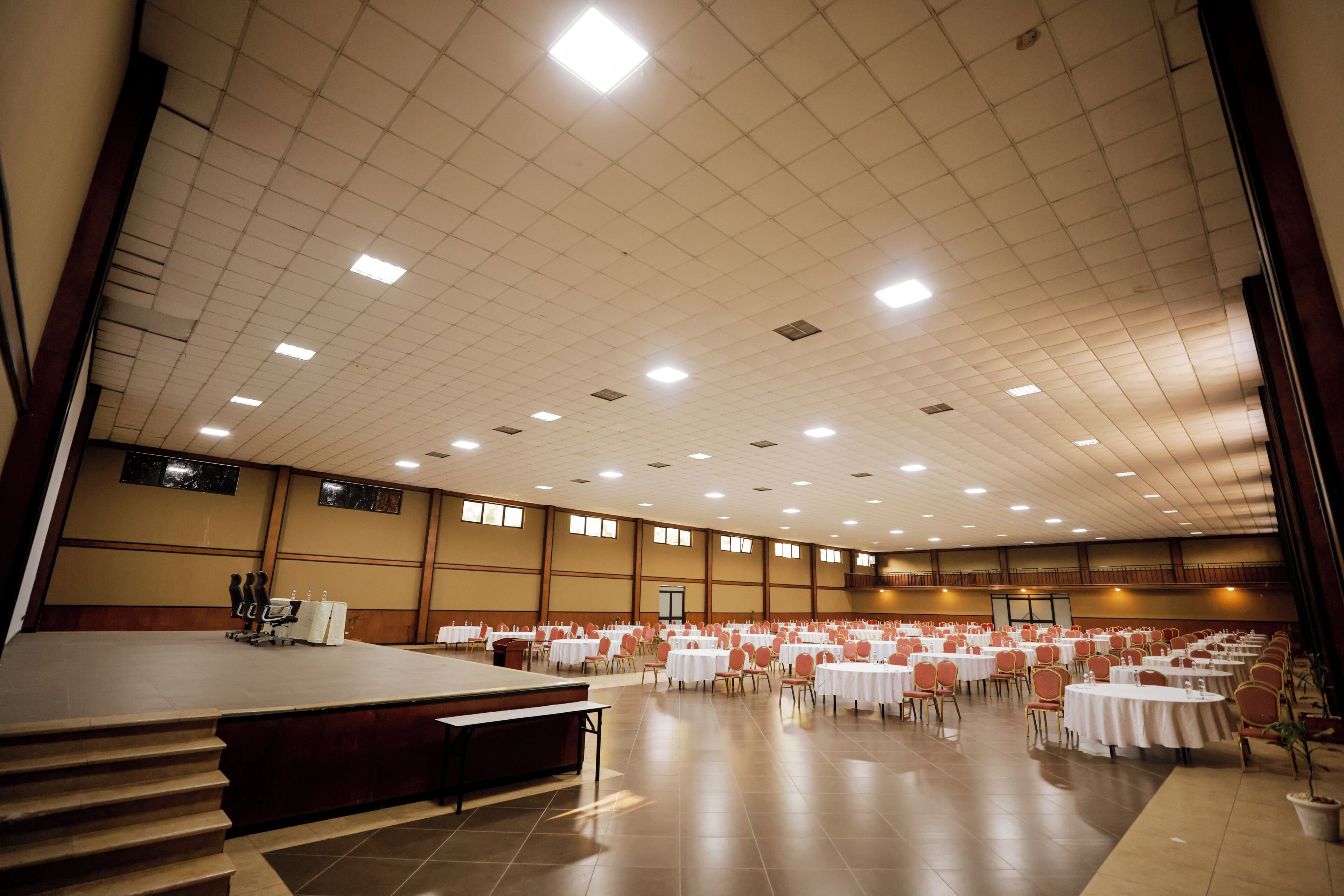 Un salón amplio con filas de mesas y sillas, un escenario y un techo alto con luces.