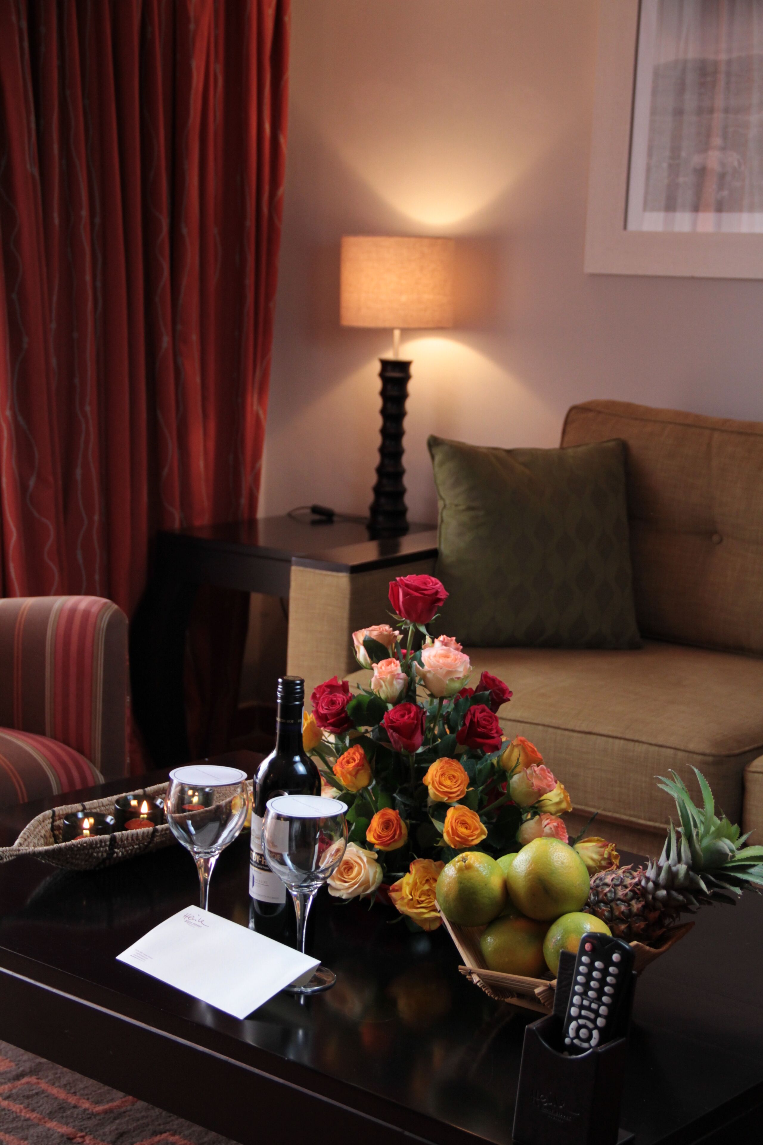 A hotel room with a dining table set with wine glasses, a remote control, a basket of fruit, and a bouquet of flowers.