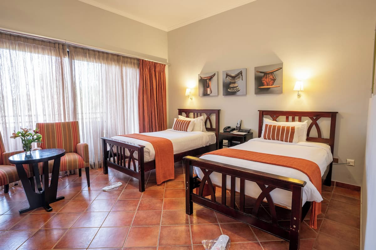 A hotel room with two beds, a small table, a chair, and a window with curtains.