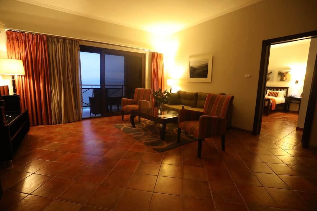 A hotel room with a living area featuring a sofa, armchairs, a coffee table, and a TV. There is a balcony with curtains, a painting on the wall, and a tiled floor.