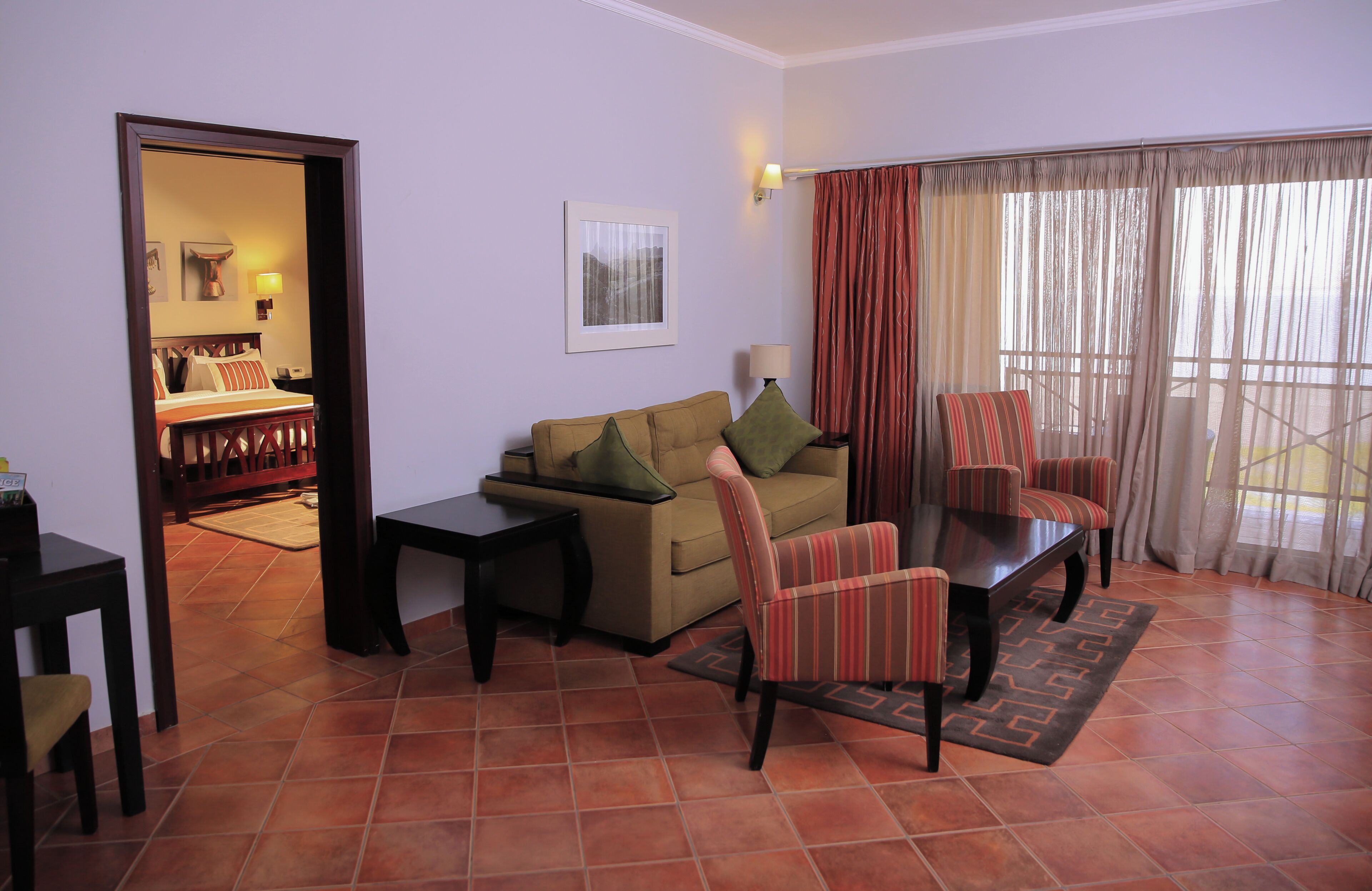 A living room with a sofa, armchairs, a coffee table, a side table, a lamp, a framed picture on the wall, and a view through the window.