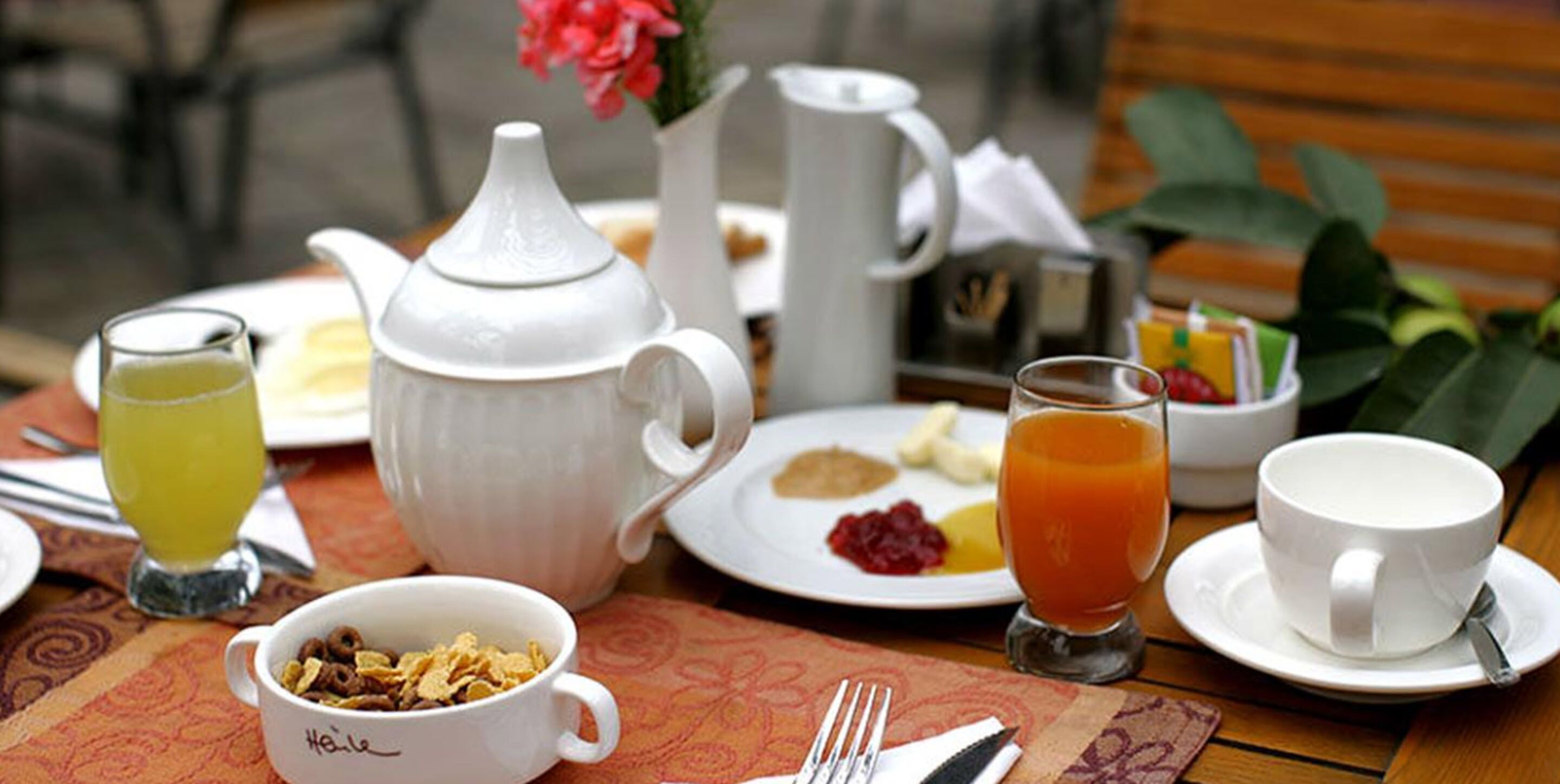 A breakfast setting with a teapot, cups, a bowl of cereal, and a glass of orange juice on a table.