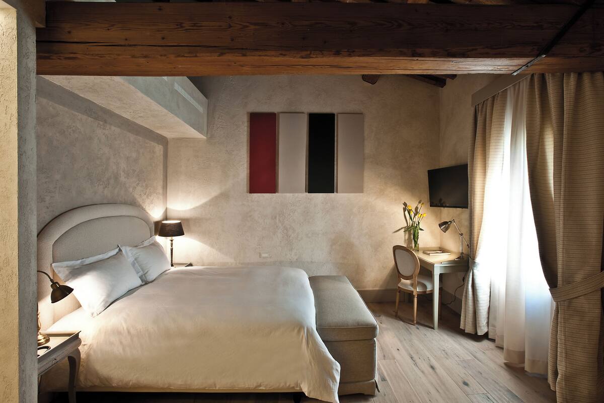Un dormitorio con cama, mesita de noche, lámpara y escritorio con silla. Hay un televisor montado en la pared y una ventana con cortinas.