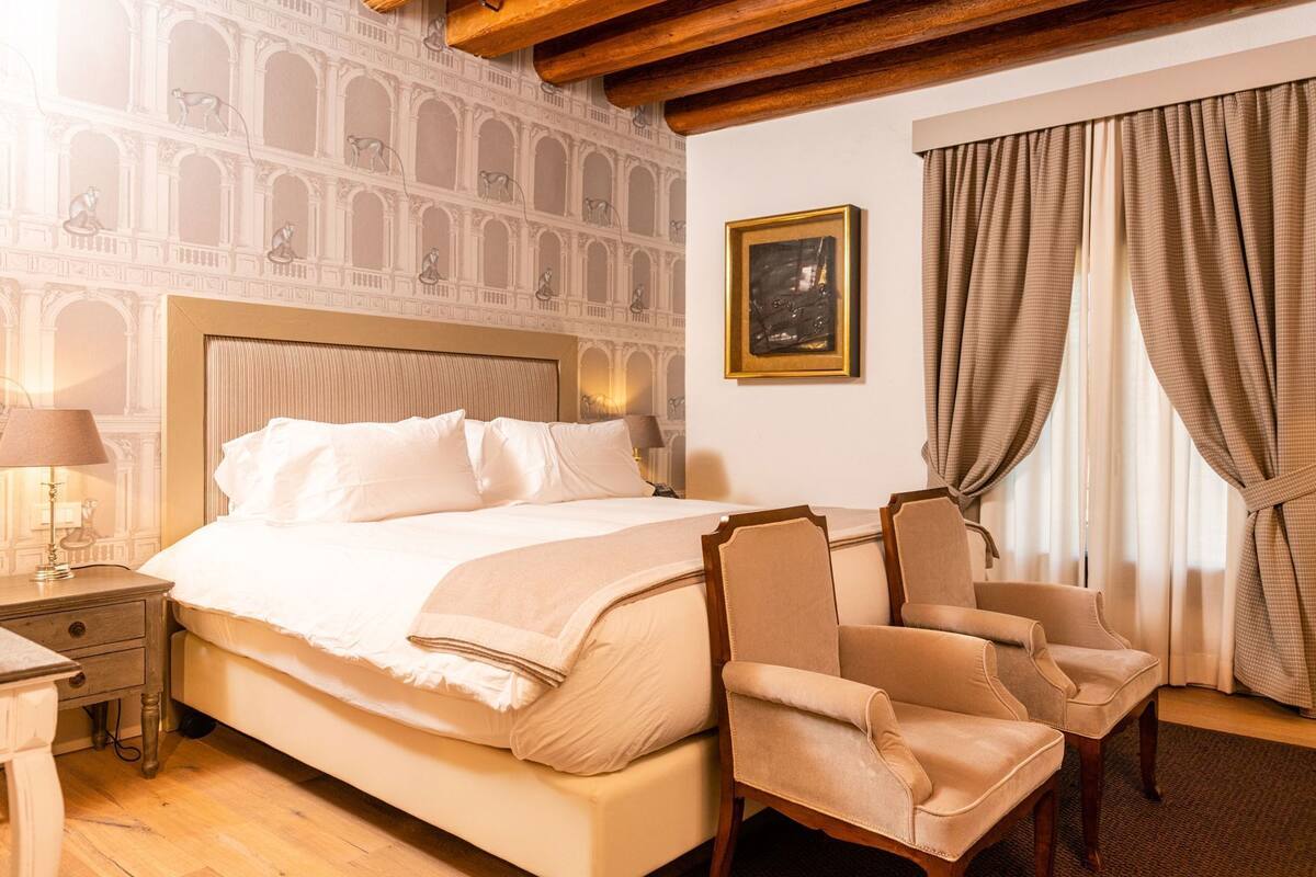 Habitación de hotel con cama, dos sillones, una mesita de noche, lámpara, cuadro enmarco y ventana con cortinas.