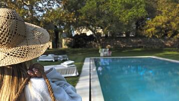 Une personne portant un chapeau de paille et tenant une serviette près d’une piscine, avec des chaises longues et une zone herbeuse en arrière-plan.