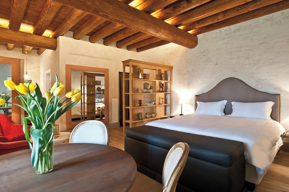 Un dormitorio con una cama grande, una estantería de madera, una mesa de comedor con sillas y un jarrón de tulipanes.