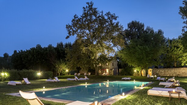 Piscina com espreguiçadeiras e velas, cercada por árvores e um muro de pedra.