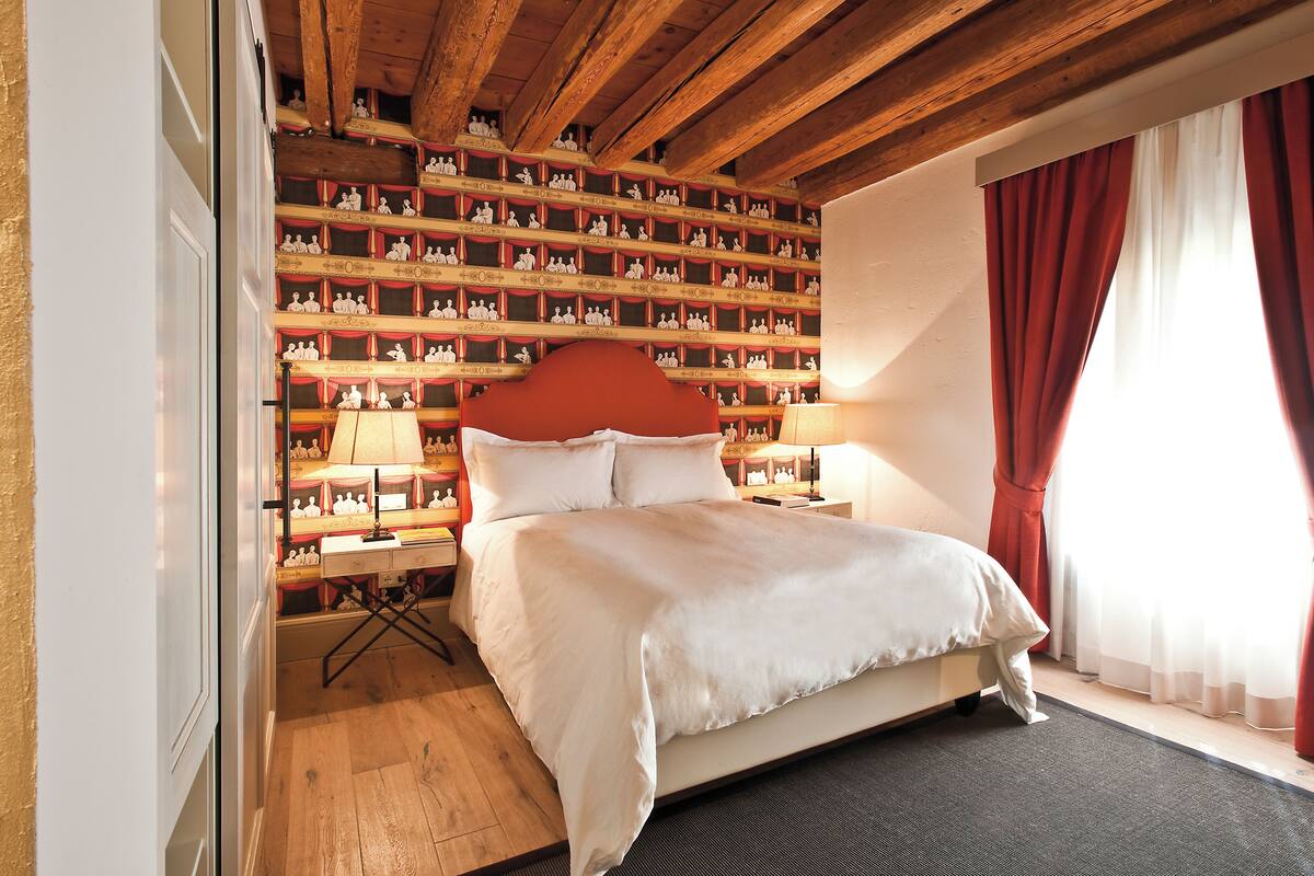 Un dormitorio con una cama grande, vigas de madera en el techo, una pared decorada con figuritas y una ventana con cortinas rojas.