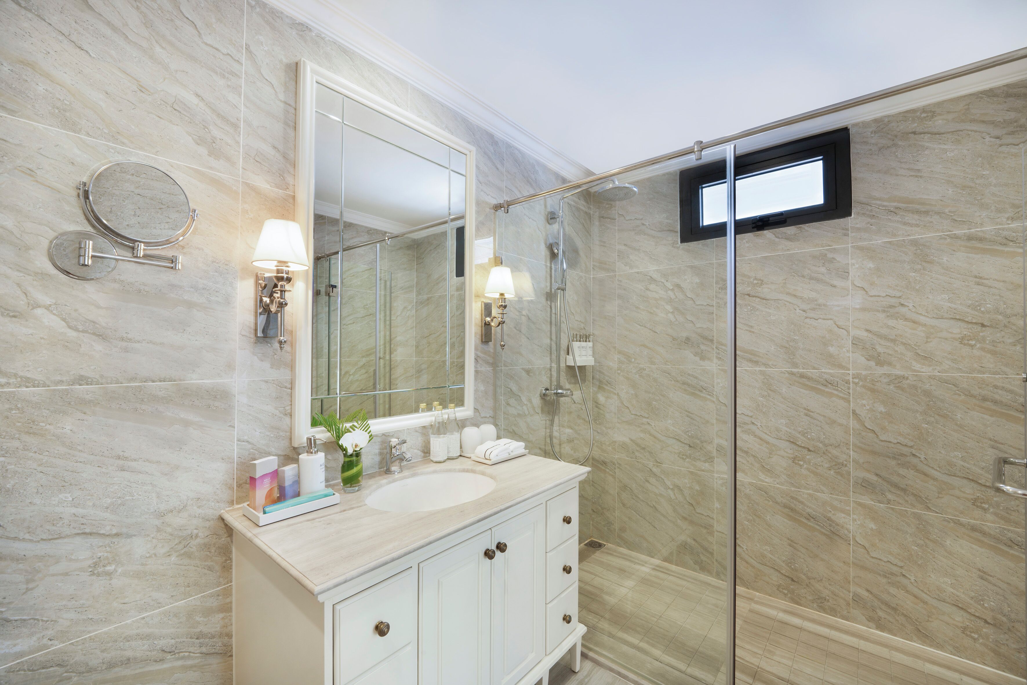 A bathroom with a marble wall, a large mirror, a sink with toiletries, and a glass shower enclosure.