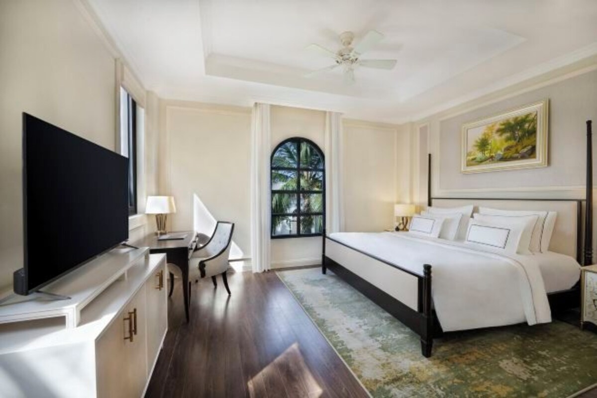Een hotelkamer met een bed, een bureau, een televisie, een raam en een plafondventilator.