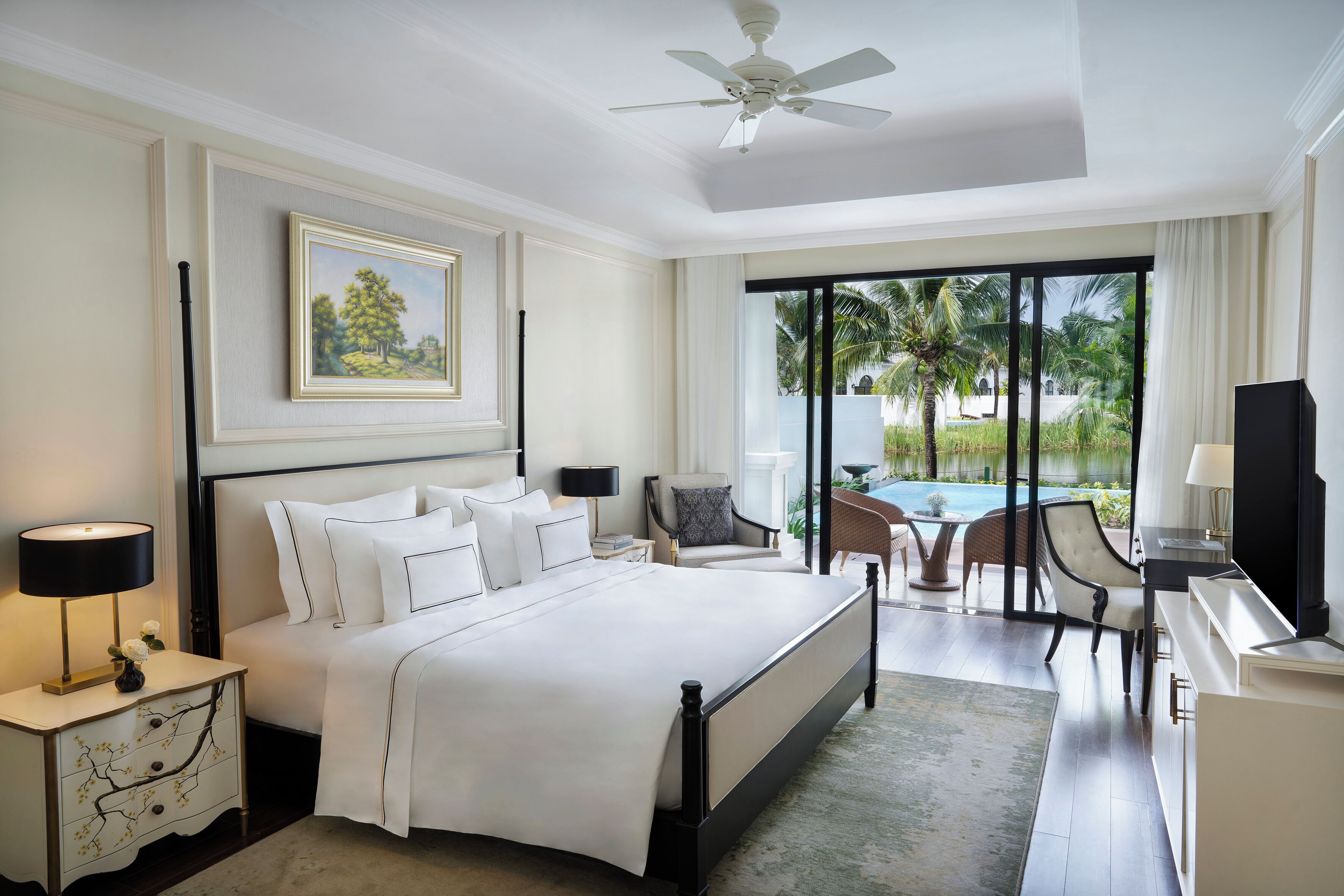 A spacious bedroom with a large bed, a desk, and a view of a pool and palm trees.