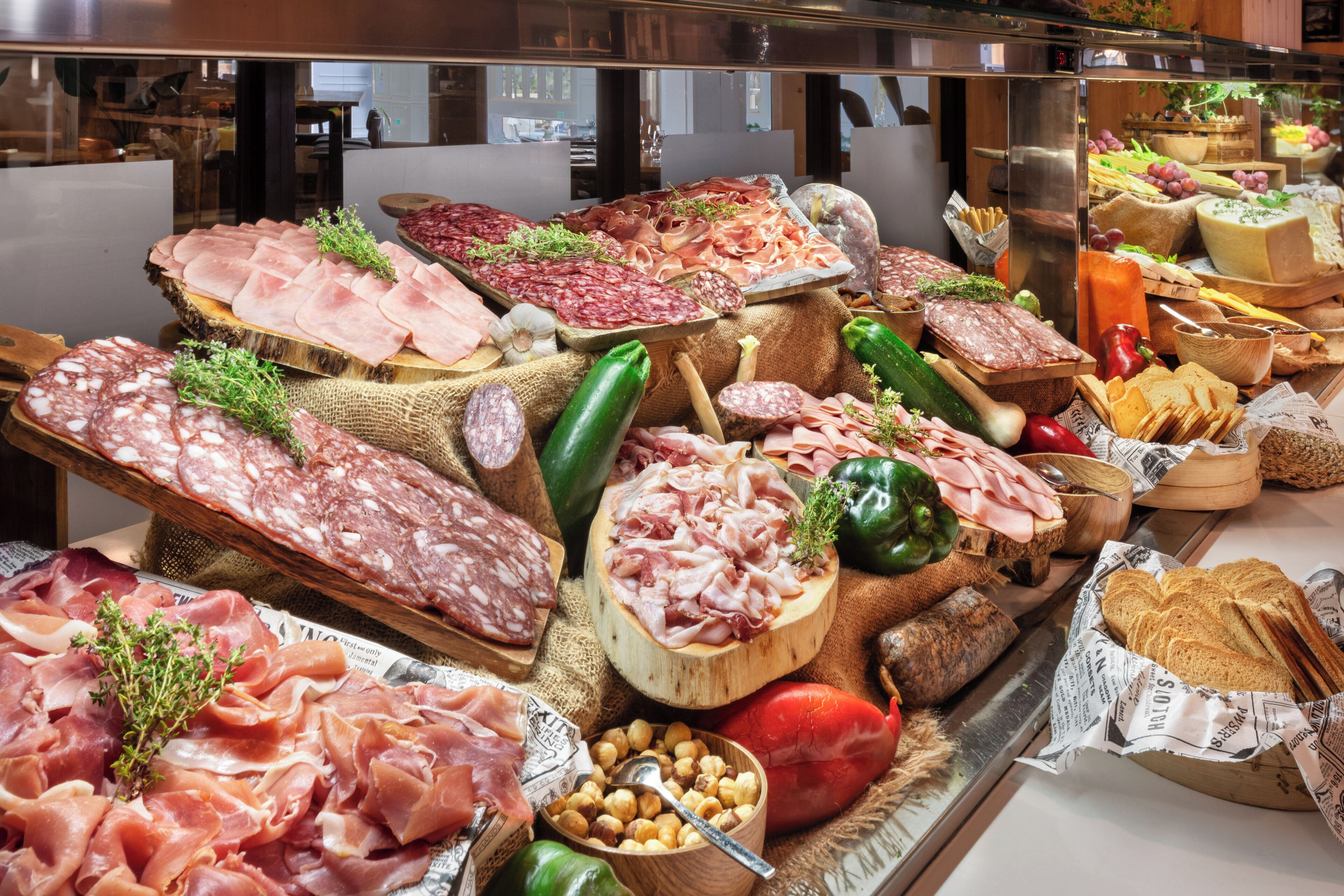 A variety of meats and cheeses displayed on a wooden board, surrounded by vegetables and bread.