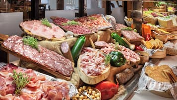 A variety of meats and cheeses displayed on a wooden board, surrounded by vegetables and bread.