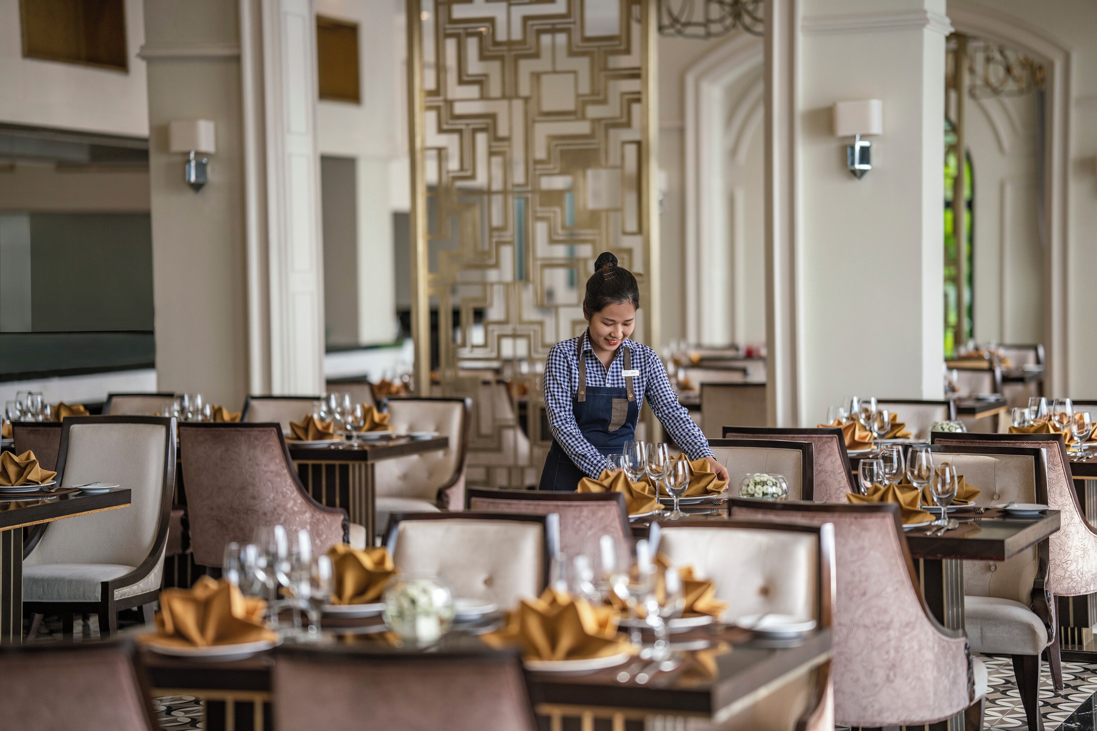 A well-appointed dining area with neatly set tables, a waitstaff attending to guests, and a decorative wall with geometric patterns.