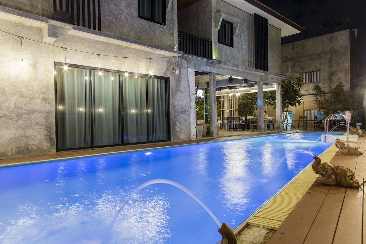 Una piscina con fuente acuática, rodeada por una zona exterior cubierta con asientos y vistas a un edificio con grandes ventanales.