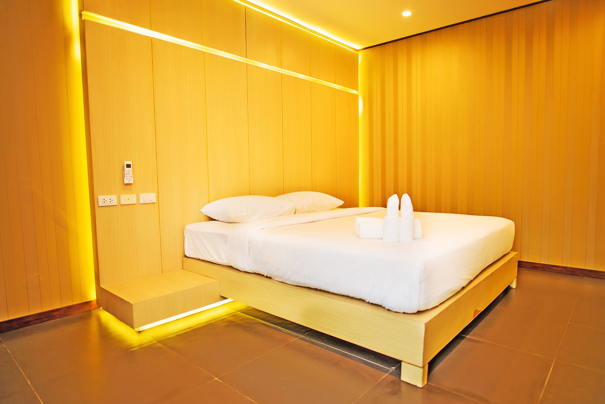 A modern hotel room with a wooden bed, bedside controls, and a small bench.