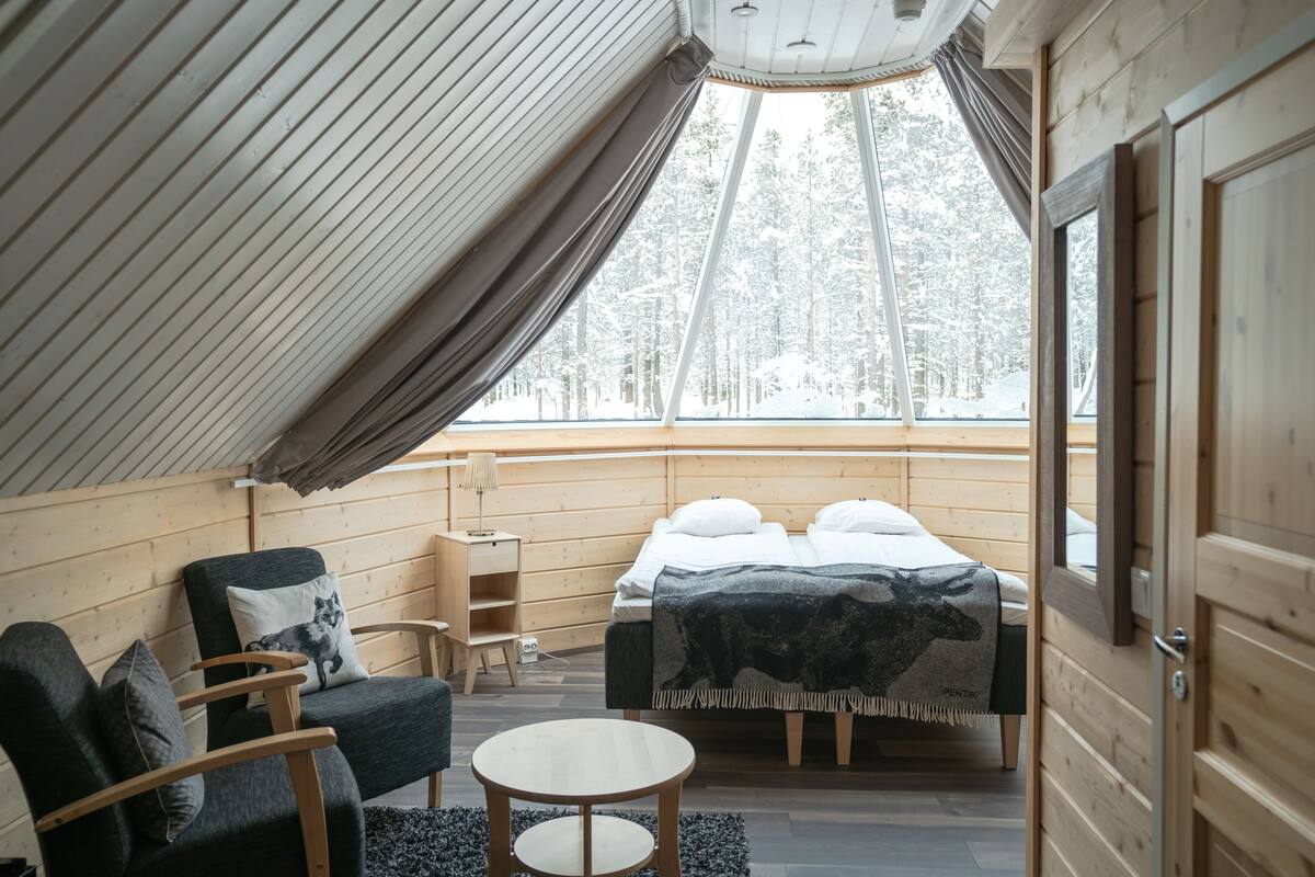 Ein gemütliches Zimmer mit einem Bett, einem Sessel, einem kleinen Tisch und einem Fenster mit Blick auf einen verschneiten Wald.