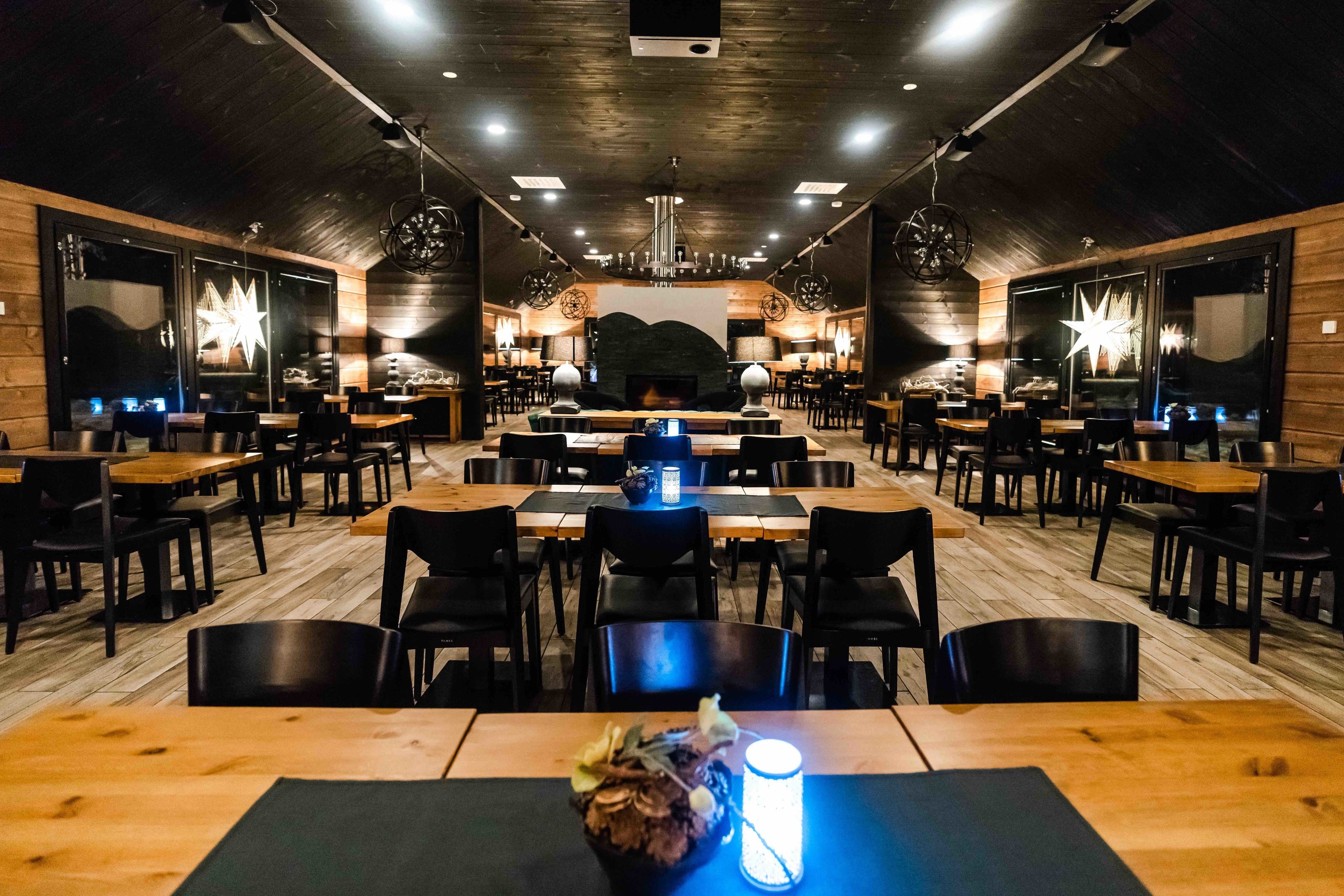 A restaurant with wooden walls and a central fireplace. Tables are arranged in rows with chairs.