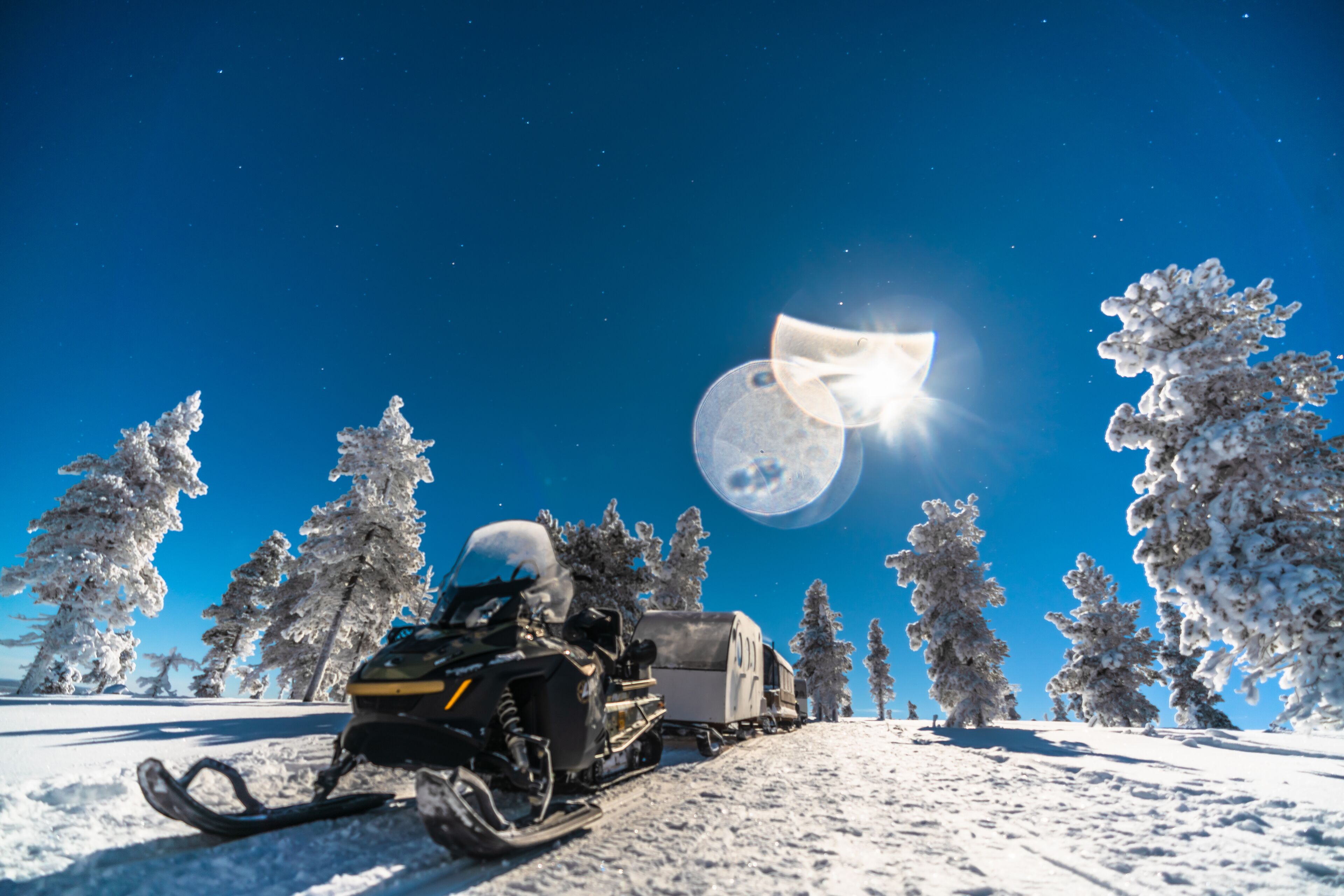 A snowmobile on a snowy path with trees, a clear sky, and a bright sun.