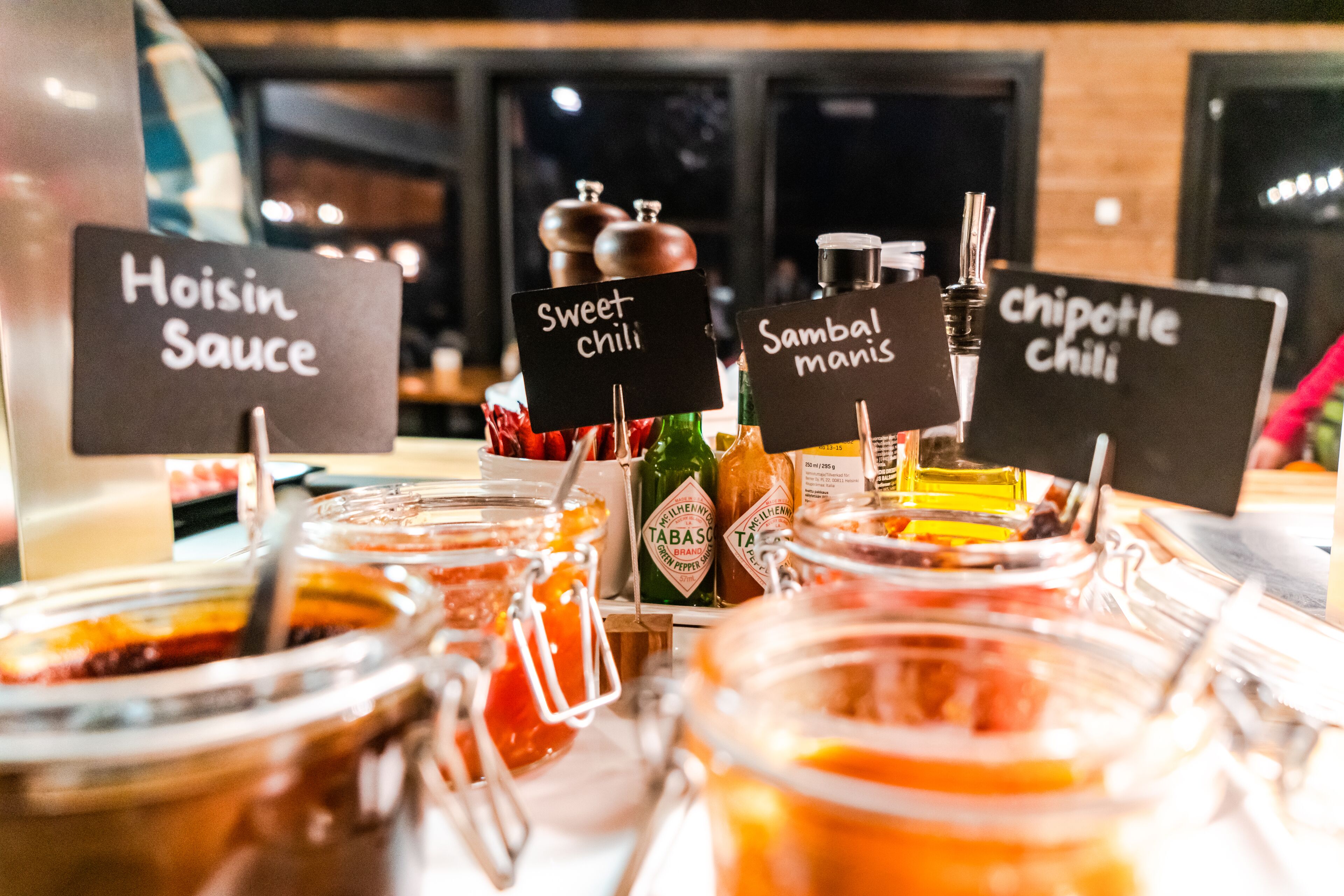 A buffet with various condiments and sauces labeled, including Hoisin Sauce, Sweet Chili, Sambal Manis, and Chipotle Chili.