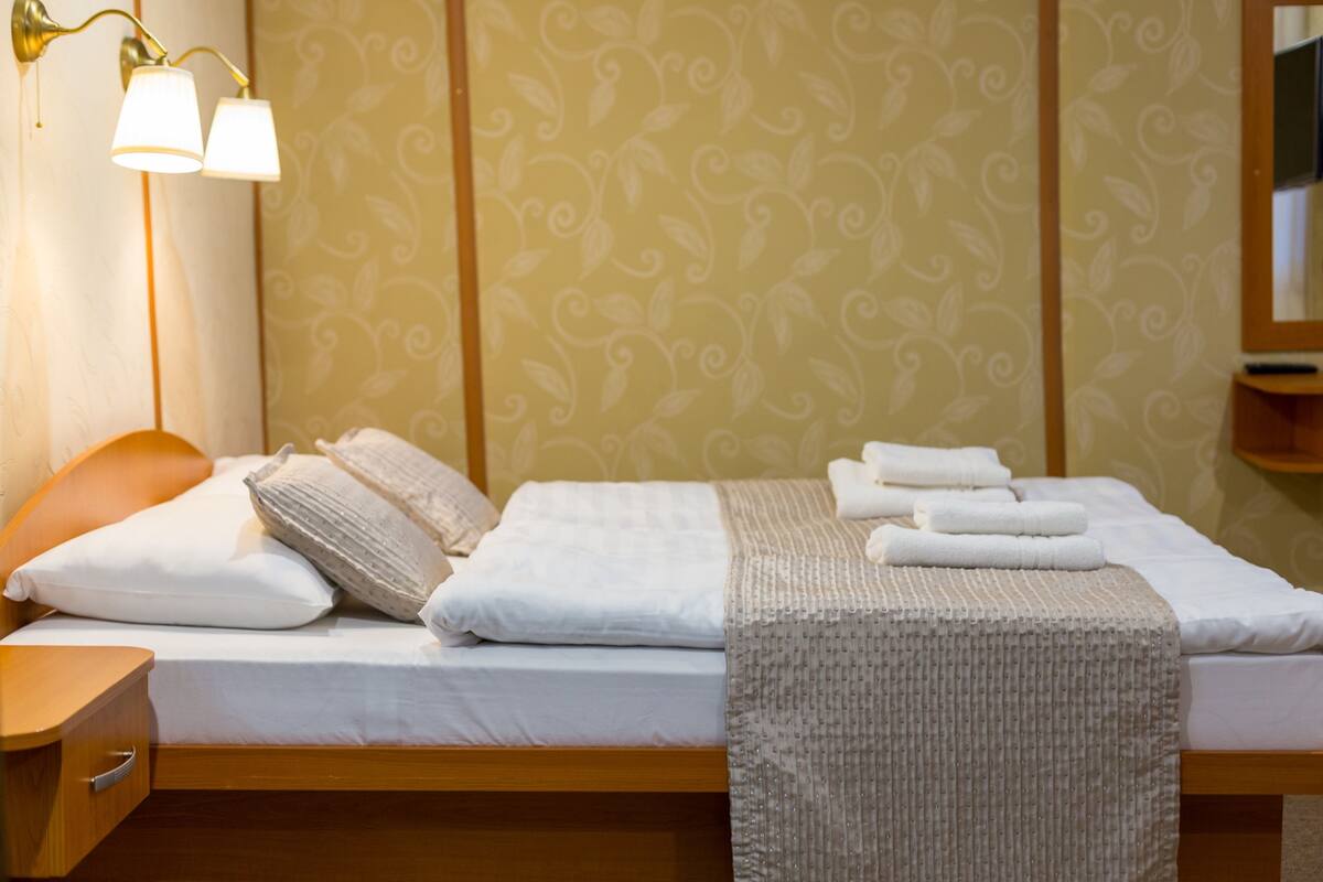 A neatly made bed with white linens, a beige throw blanket, and a set of towels. There is a bedside table with a lamp and a wooden nightstand. The wall features a patterned wallpaper with a leaf design.