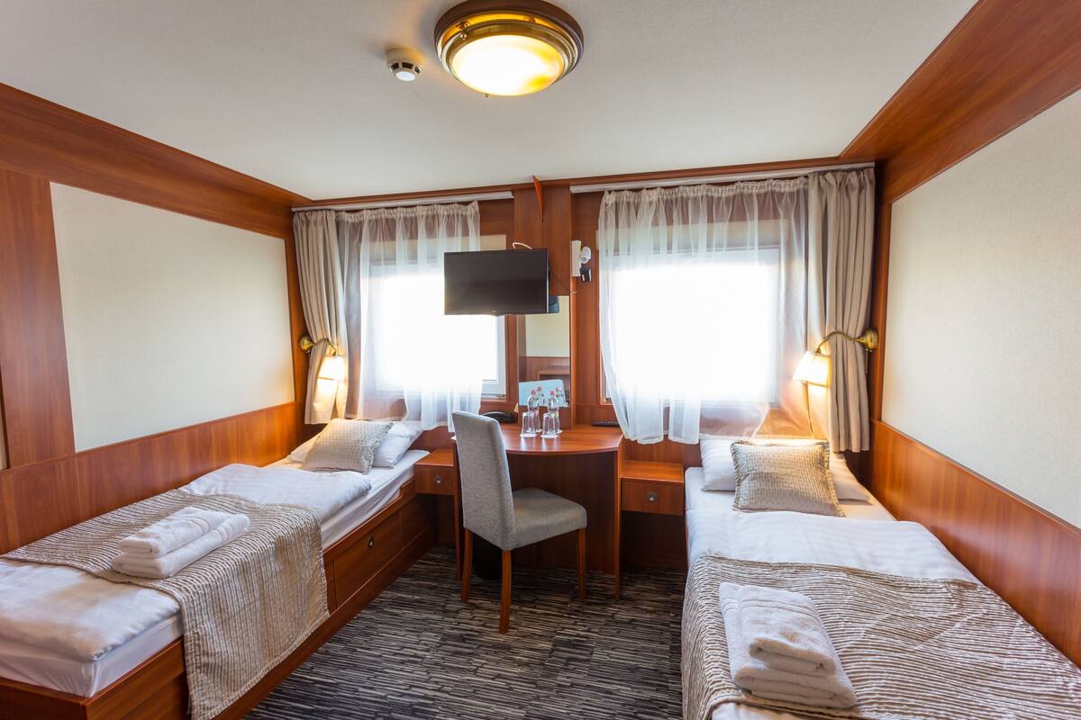 A hotel room with two single beds, a desk with a chair, a television, and a window with curtains.