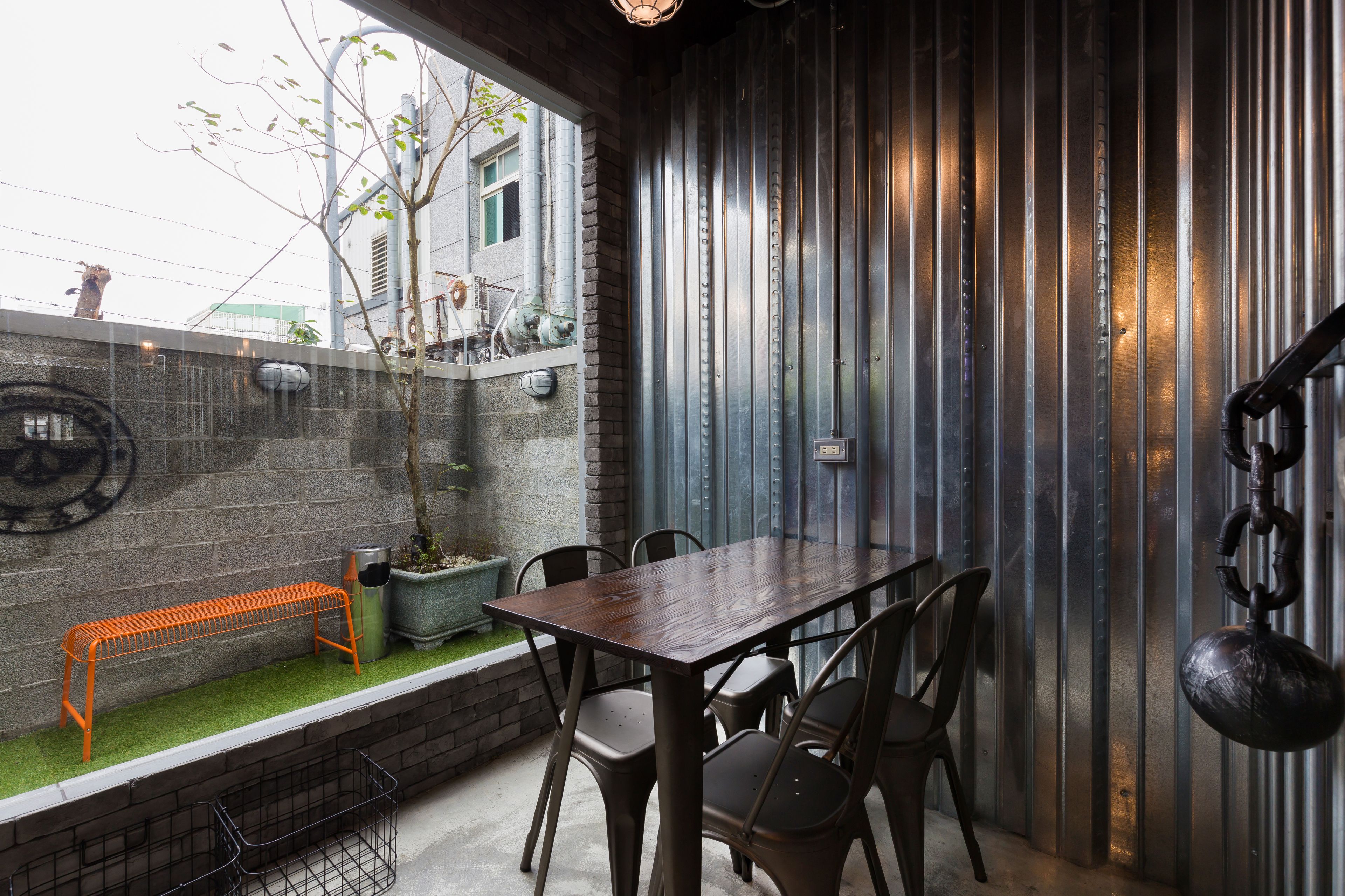 A patio area with a table and chairs, a metal bench, a potted plant, and a view of a building through a window.