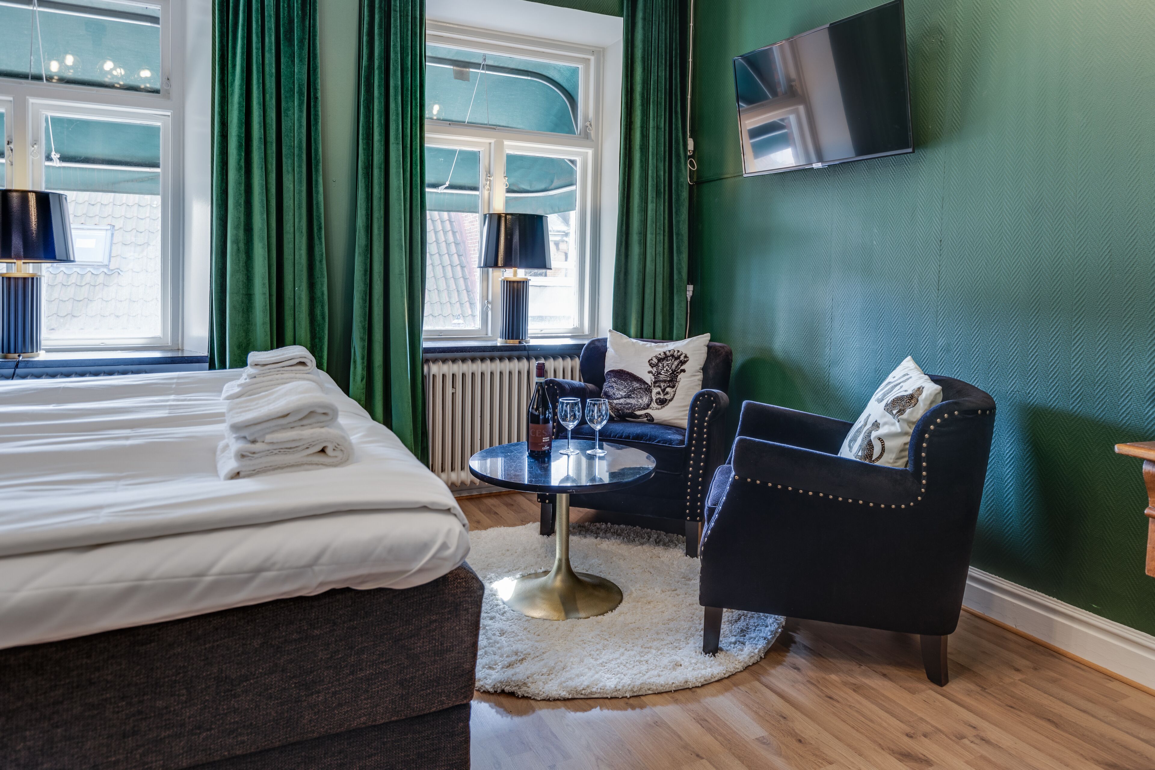 Una habitación de hotel con una cama bien hecha, una mesa redonda con dos copas de vino, un sillón negro y un televisor montado en la pared.