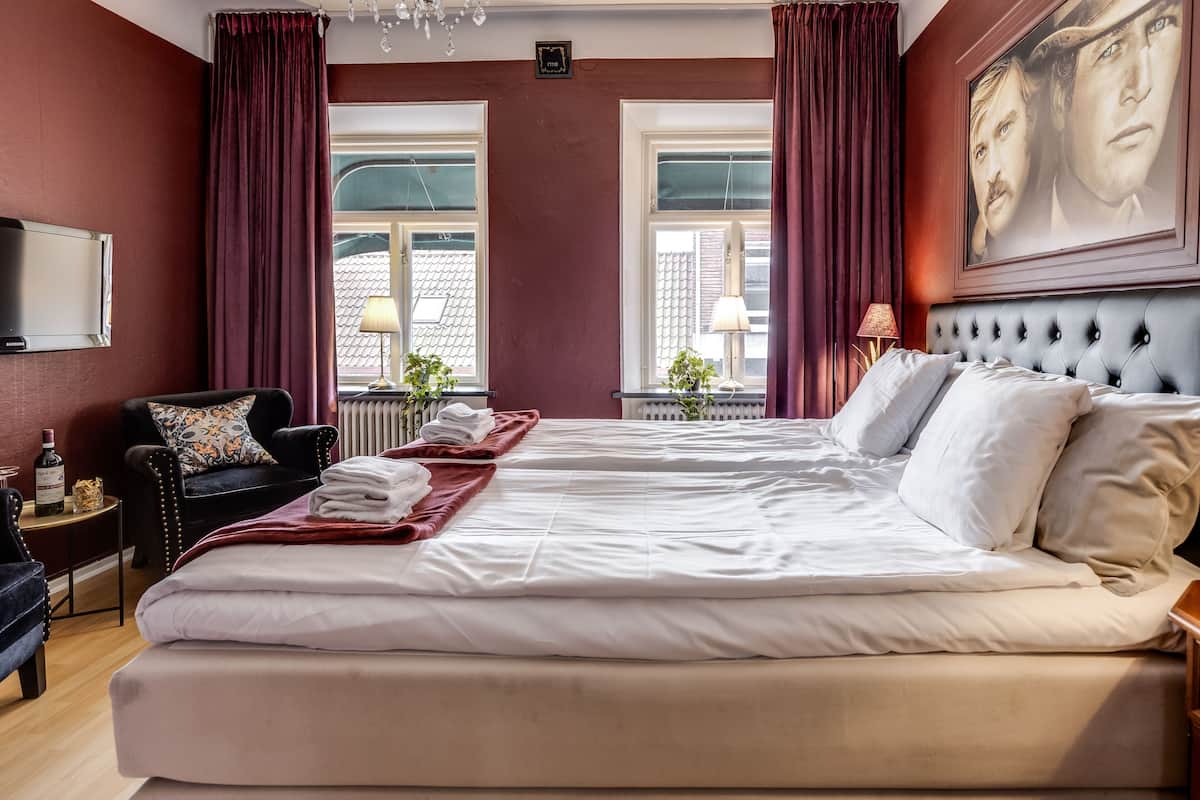Une chambre d’hôtel avec un grand lit, deux fenêtres avec des rideaux, un fauteuil noir, une petite table avec une lampe et un tableau au mur.