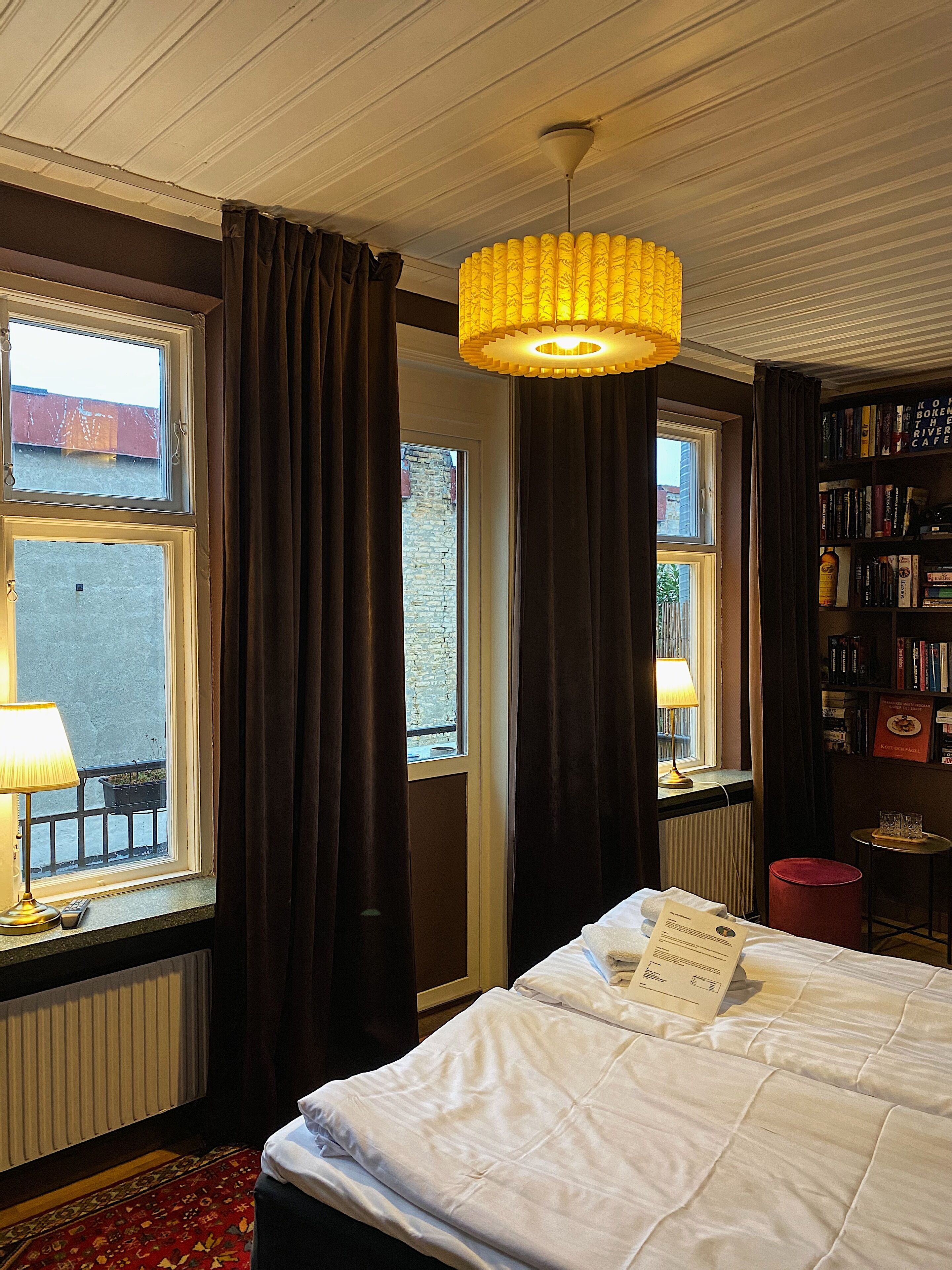 Una habitación de hotel con una cama, una ventana con cortinas, una estantería y un candelabro.