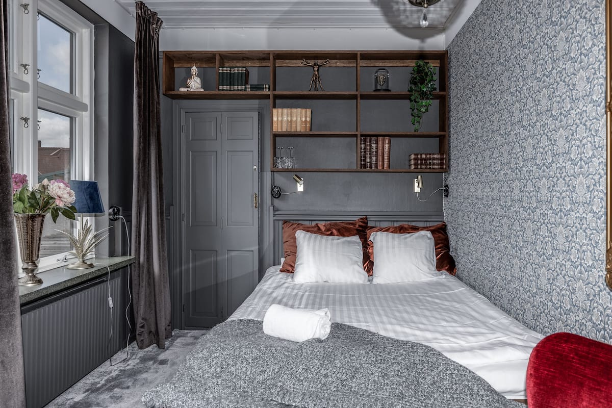 Une chambre à coucher bien rangée, avec un lit, une table de chevet dotée d’une lampe, une étagère avec des livres et des objets décoratifs, et une fenêtre avec des rideaux.