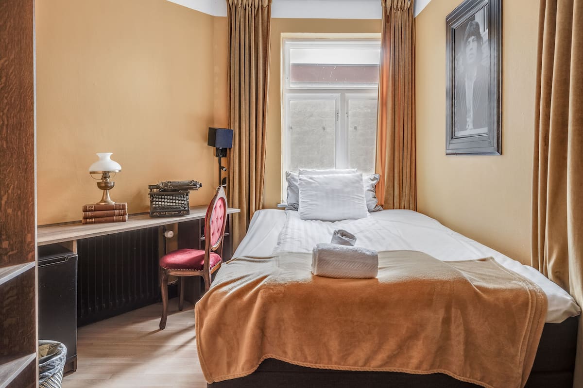 Une chambre d’hôtel avec un lit, un bureau avec une lampe, une chaise et une fenêtre avec des rideaux.