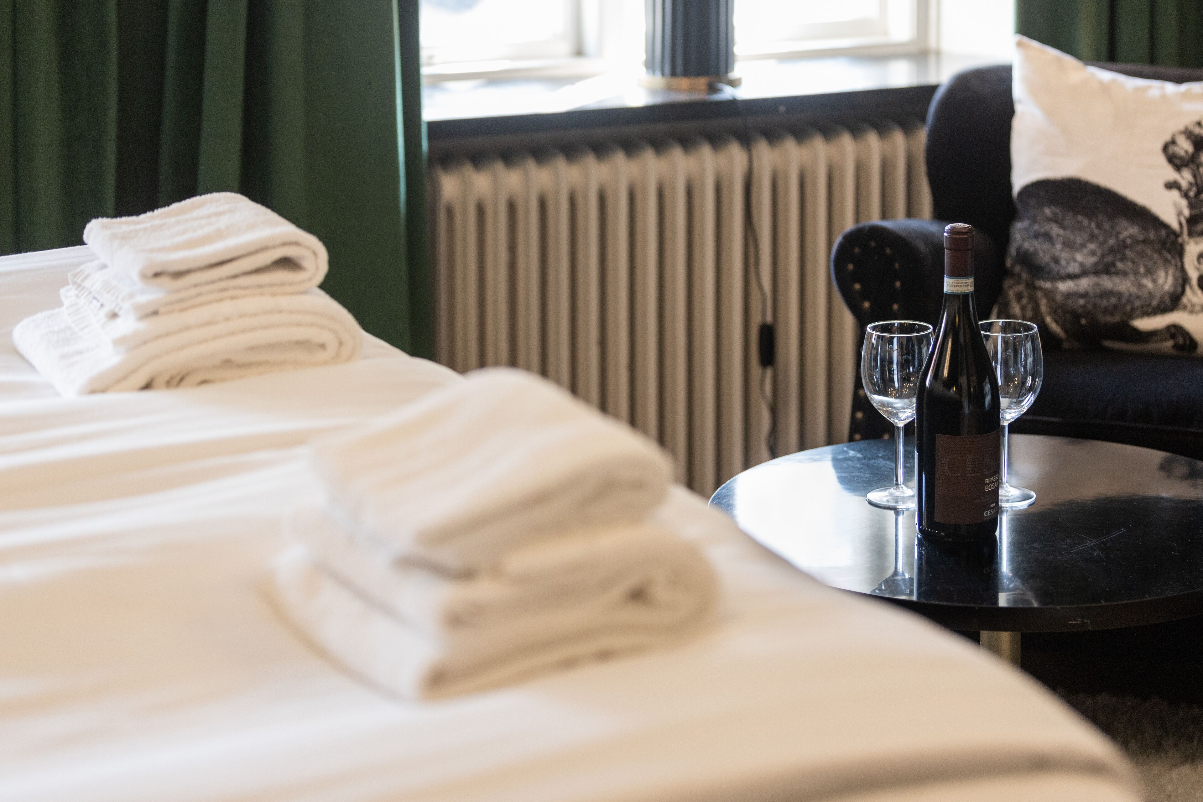 Una cama bien hecha con toallas blancas dobladas, una botella de vino y dos copas sobre una mesa redonda en una habitación de hotel.