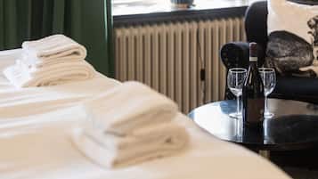 Una cama bien hecha con toallas blancas dobladas, una botella de vino y dos copas sobre una mesa redonda en una habitación de hotel.