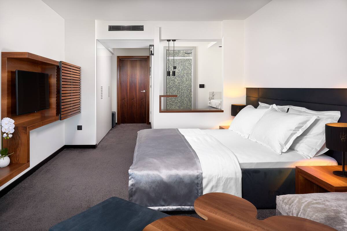 A modern hotel room with a large bed, a TV mounted on a wooden panel, a shower area, and a small seating area.
