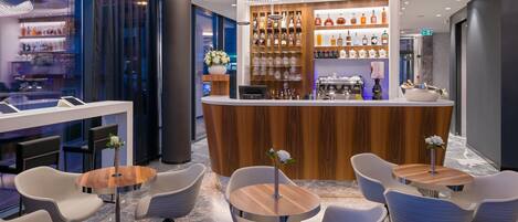 Eine moderne Bar mit Holztresen, verschiedenen Spirituosenflaschen und einer Sitzecke mit weißen Stühlen und runden Tischen.