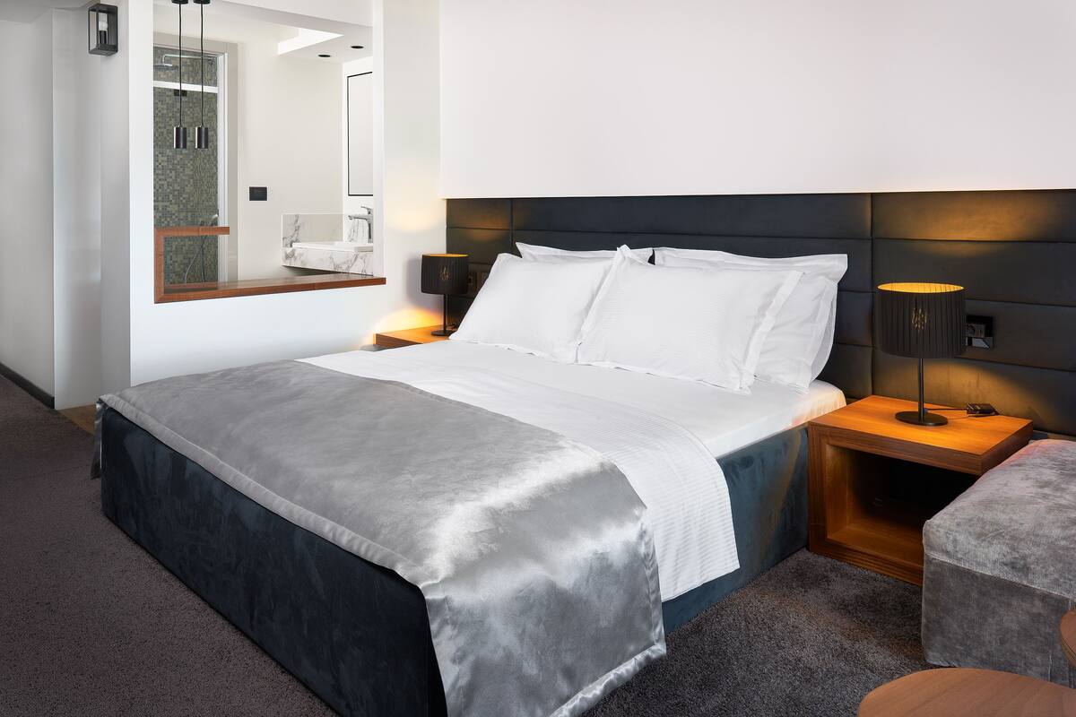 A modern hotel room with a large bed, a nightstand with a lamp, a wooden headboard, and a mirror above the bed.