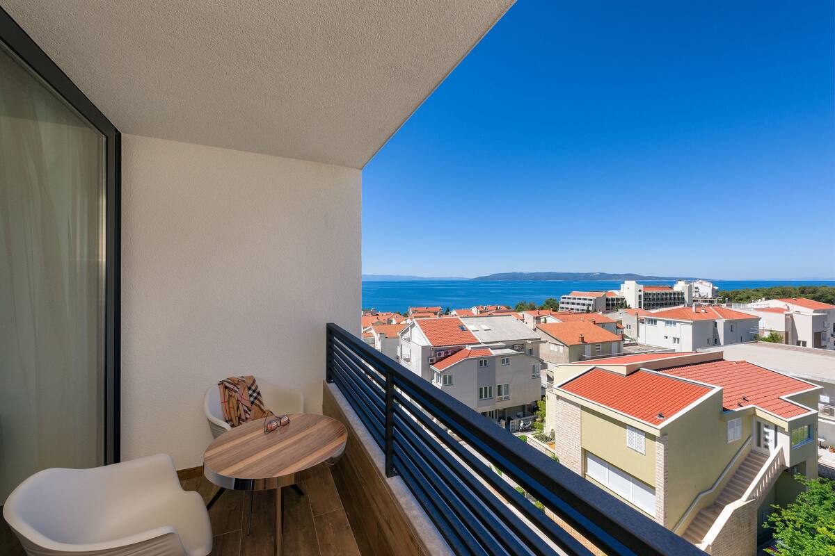 A balcony with a wooden table and white chair, overlooking a coastal town with red-roofed buildings.