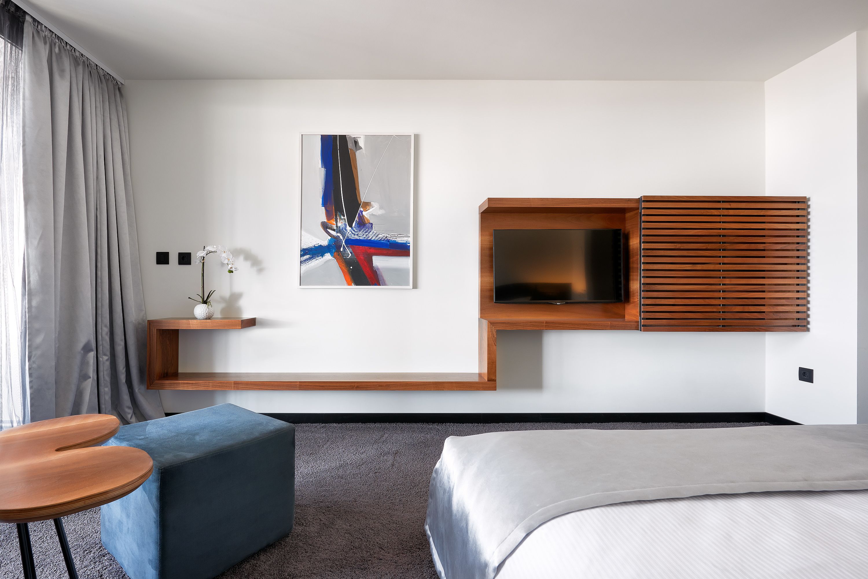 Una habitación de hotel moderna con una cama, un sofá azul, una mesita pequeña, un televisor montado en un panel de madera y un cuadro en la pared.